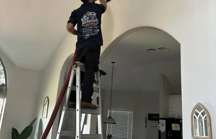 Air Busters technician on a ladder cleaning a ceiling air vent in DFW